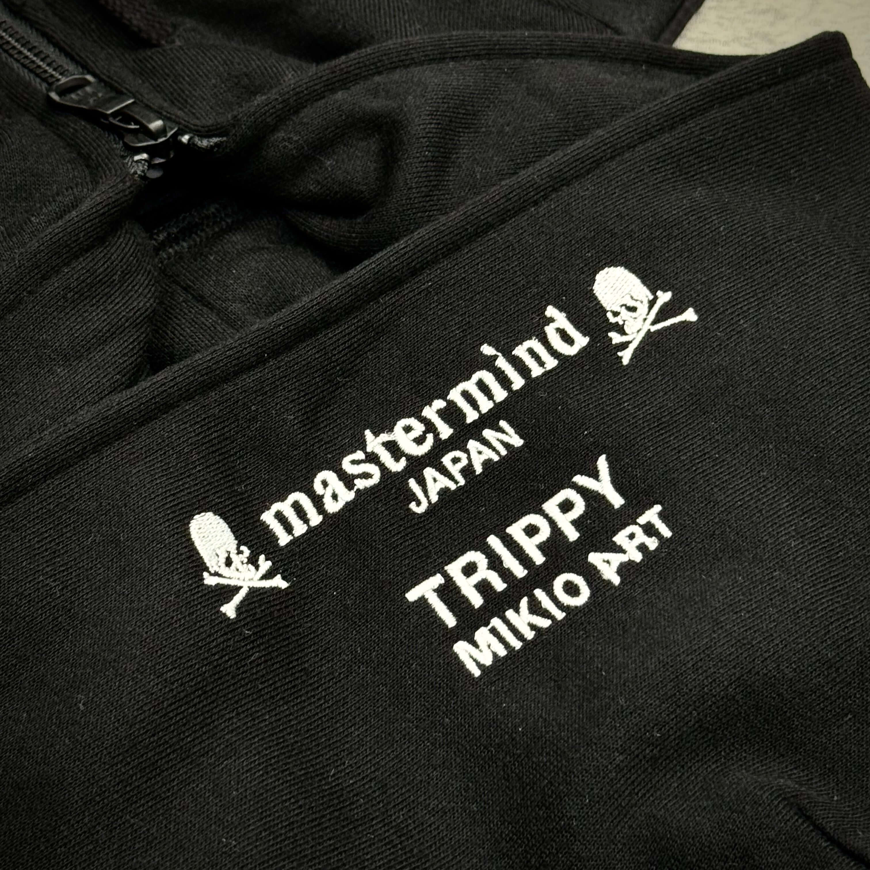 Close-up of Mastermind Japan x Trippy Art logo on a black zip-up hoodie, showcasing distinct design details.