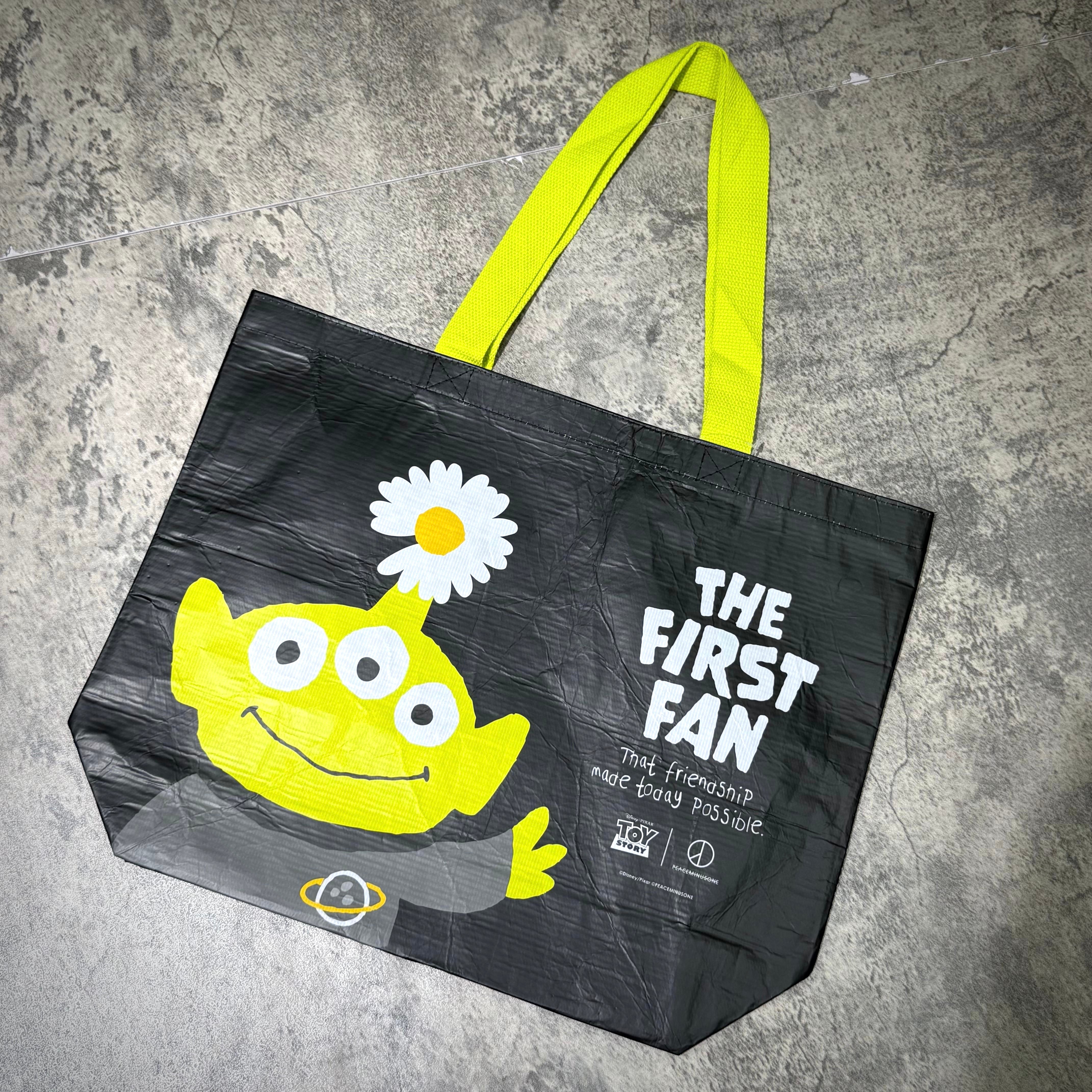 PEACEMINUSONE x Toy Story reusable bag featuring yellow alien character with text 'THE FIRST FAN' and daisy design.