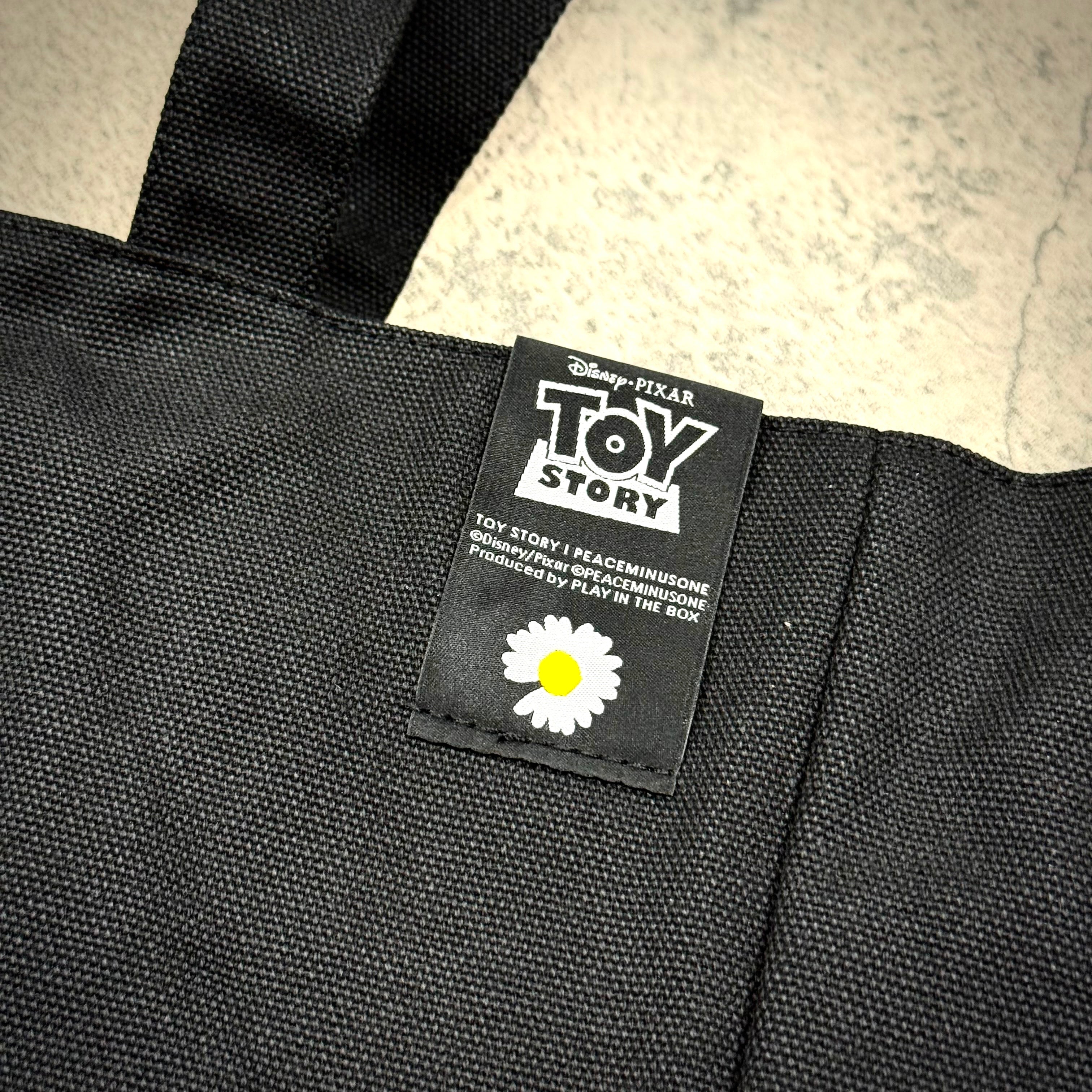 Close-up of the label on PEACEMINUSONE x Toy Story The First Fan tote bag featuring Toy Story branding and a daisy graphic.