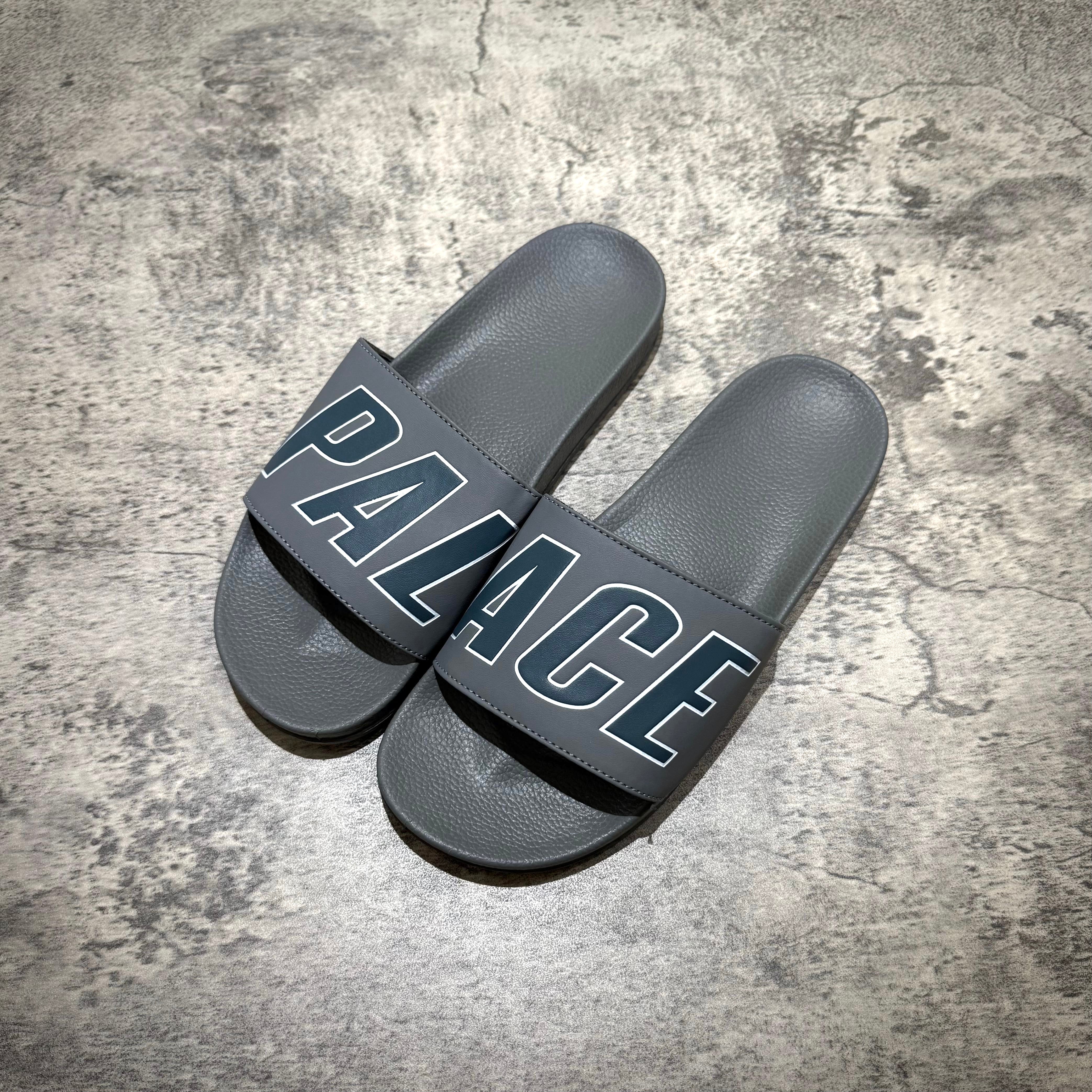 Palace Slider - Navy / Grey footwear with bold 'PALACE' print on strap, laid flat on textured background.