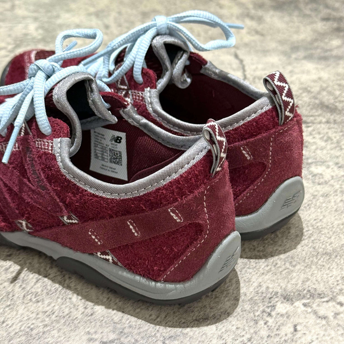 Close-up view of the backside of SUNLOVETOUR x New Balance MT1001ND shoes featuring burgundy color and light blue laces.