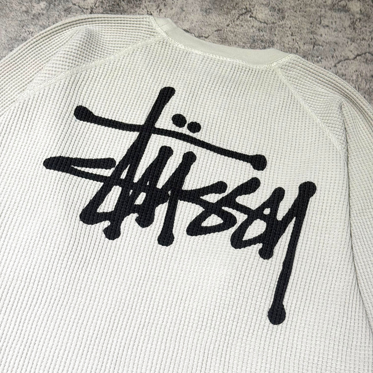 Stussy Basic Raglan Thermal LS Crew FW26 featuring iconic logo on the back with a textured white backdrop.