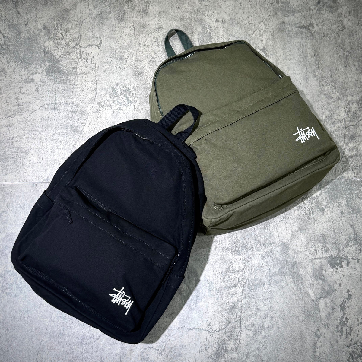 Stussy Canvas Backpack available in black and olive green color options on a textured background.