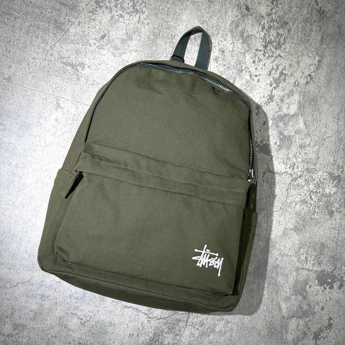 Stussy Canvas Backpack in olive green with signature logo, showcasing a sleek design on a textured background.