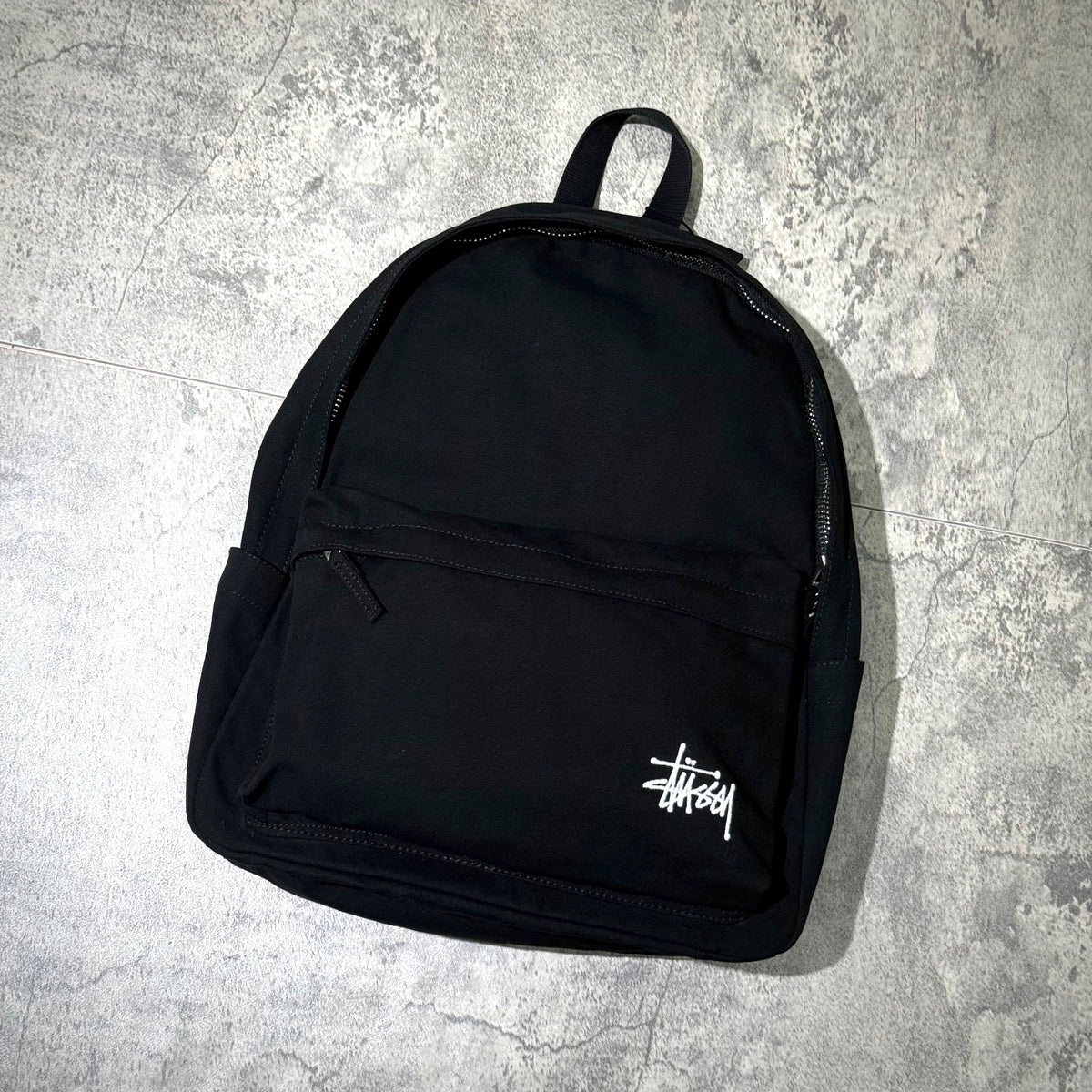 Stussy Canvas Backpack featuring a sleek black design with a front pocket and Stussy logo.