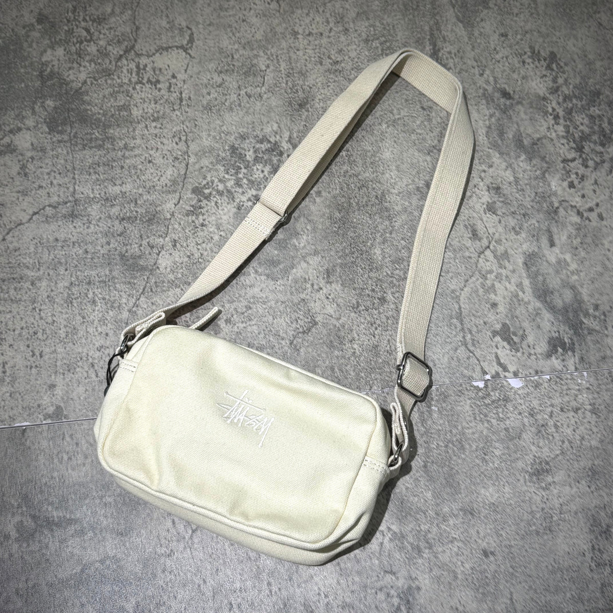 Stussy Canvas Side Pouch on a textured surface, showcasing a neutral color and adjustable strap.