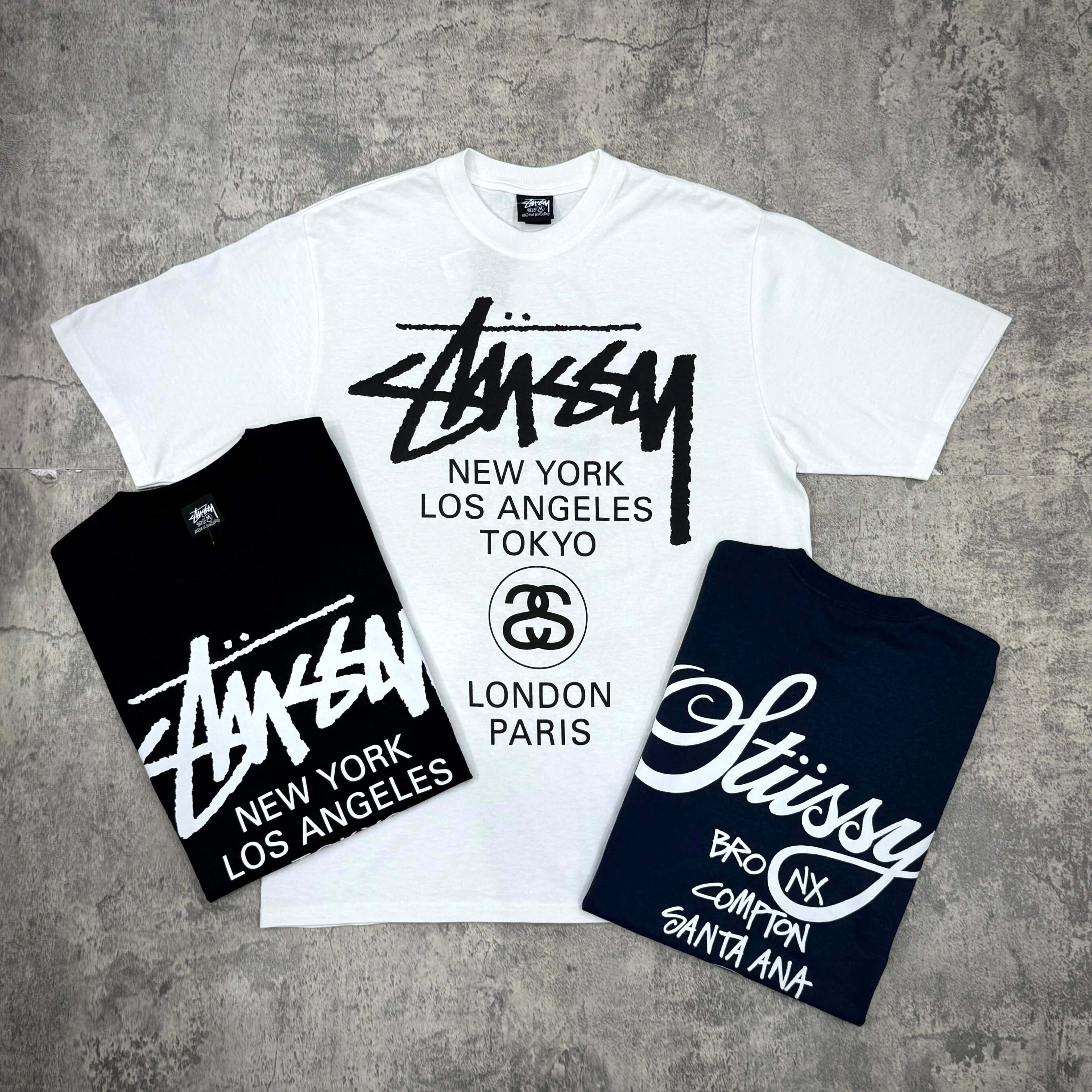 現貨 Stussy World Tour Tee - Navy / Black / White，包含三件不同顏色的Stussy T恤，顯示品牌標誌及城市名稱