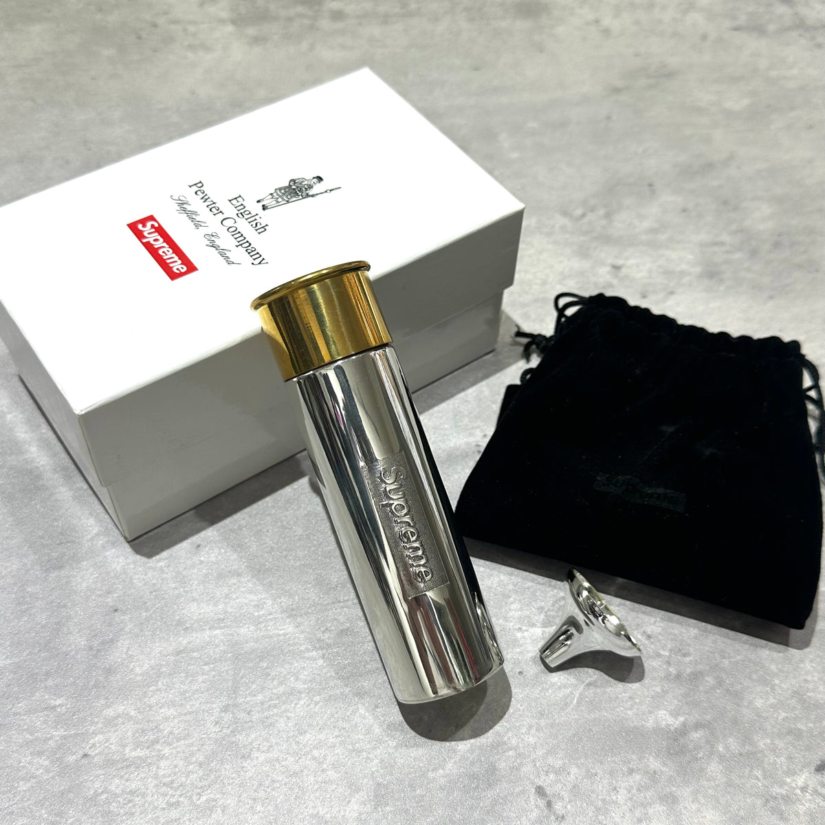 Supreme English Pewter Company Bullet Flask next to its box and accessories on a gray surface.