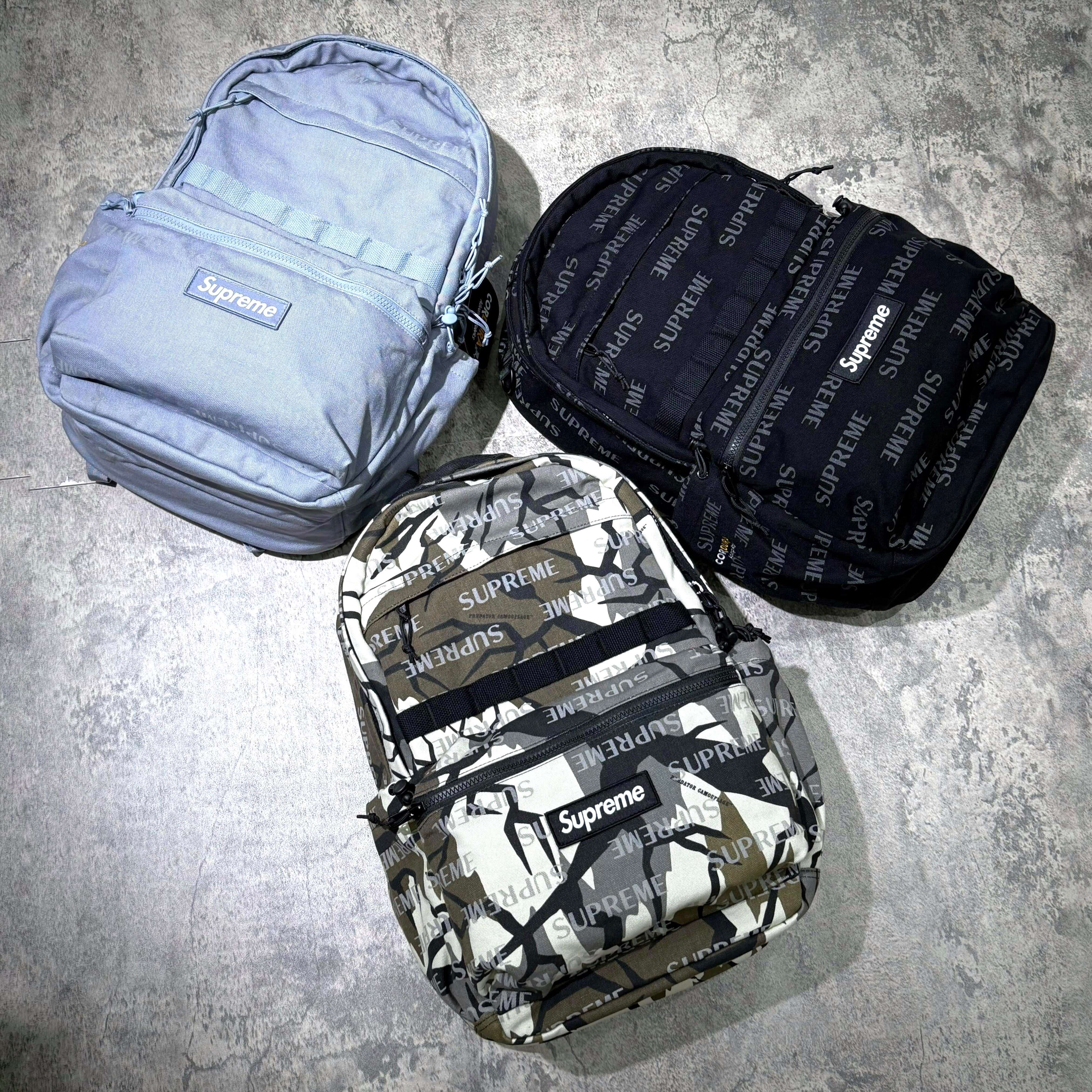 Supreme FW26 Backpack in blue, black, and camo colors displayed on a concrete surface.