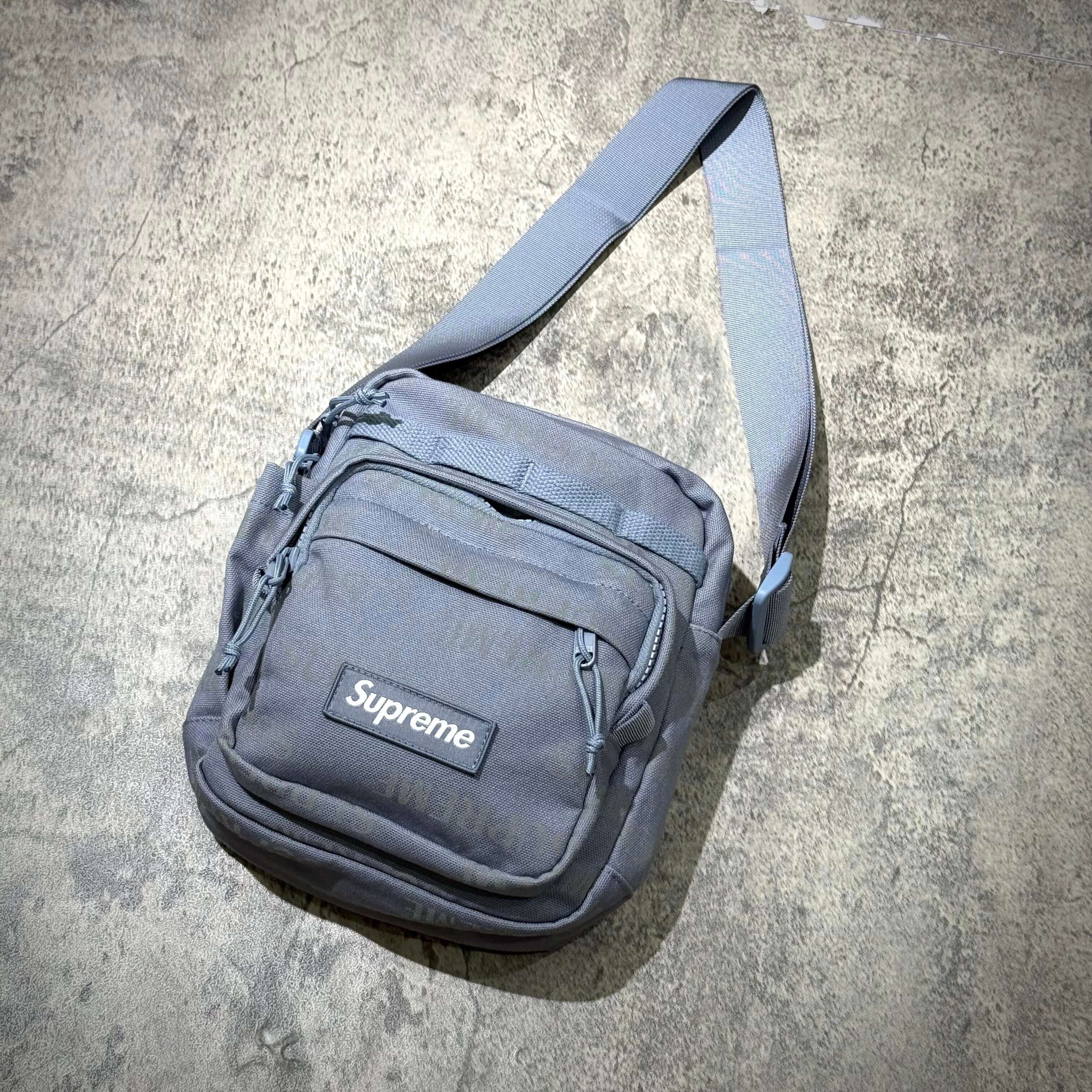 Supreme FW26 Shoulder Bag in gray with adjustable strap and front zip pocket on concrete background.