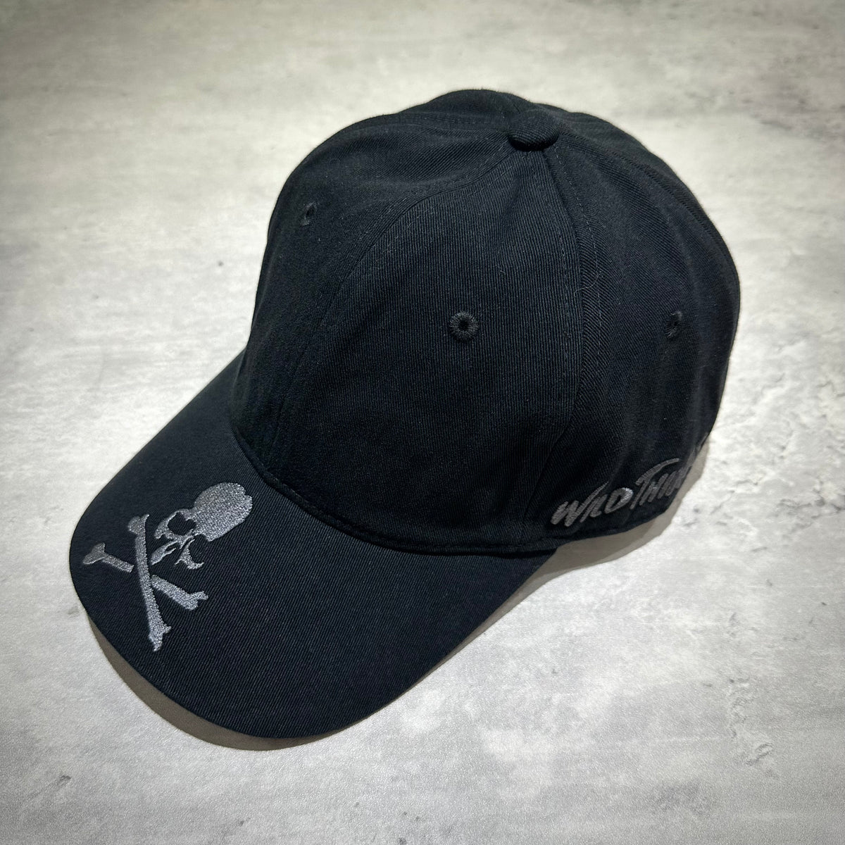 Wild Things x Mastermind World Tee Cap with embroidered skull and crossbones design on a black baseball cap.
