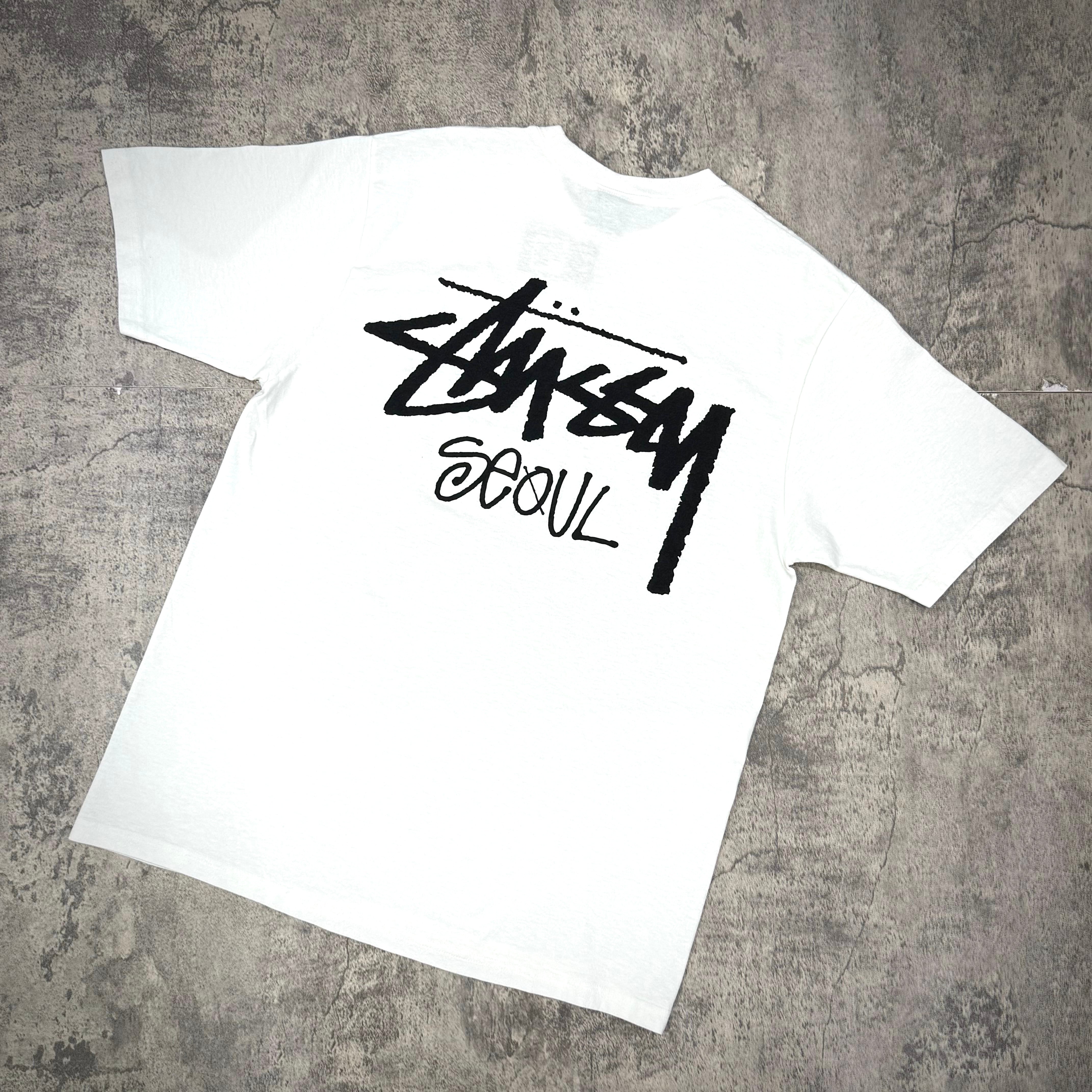 Stussy Seoul Exclusive Logo Tee in white with bold black logo on a textured background.